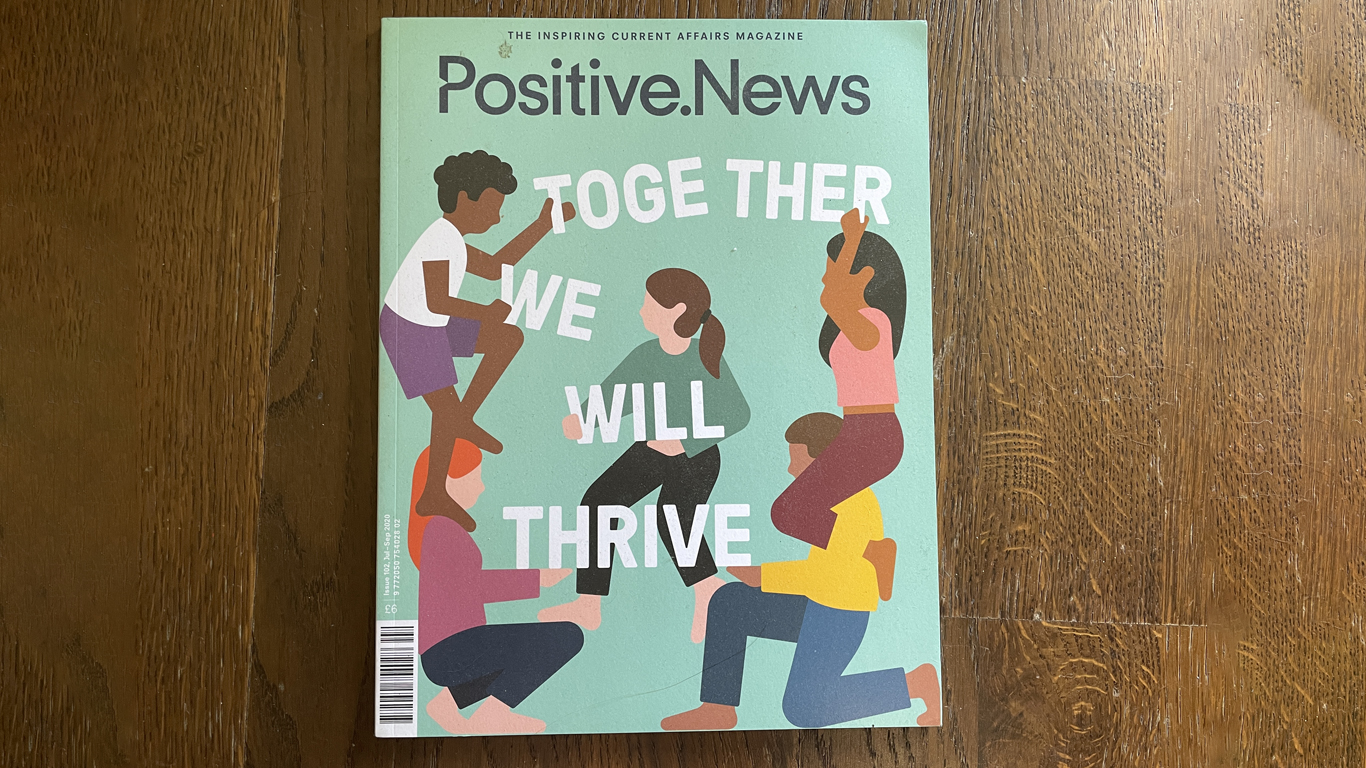 a photograph of the magazine 'Positive News'. The cover illustration shows people building words that say 'Together we will thrive' 